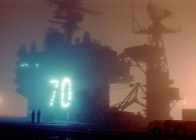 This photo shows the number 70 illuminated through the fog on the deck of an aircraft carrier behind two men.